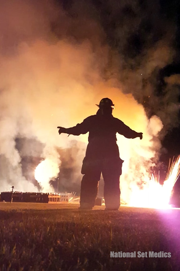 Silhouette of a firefighter with arms outstretched against fireworks and flames at night