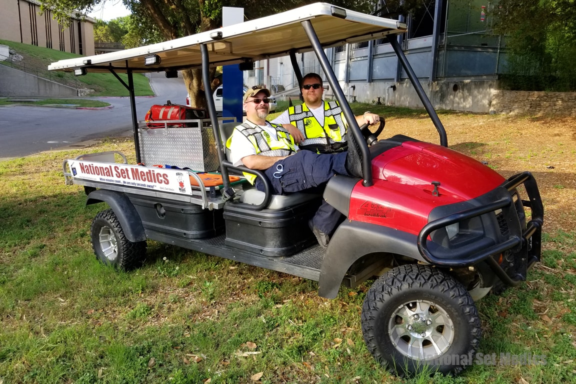 Two NSM medics in safety vests seated in a branded National Set Medics golf cart